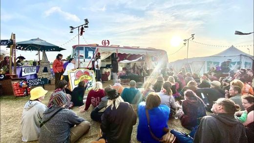 A crowd sitting on the grass at sunset watching an act in front of The Love Bus